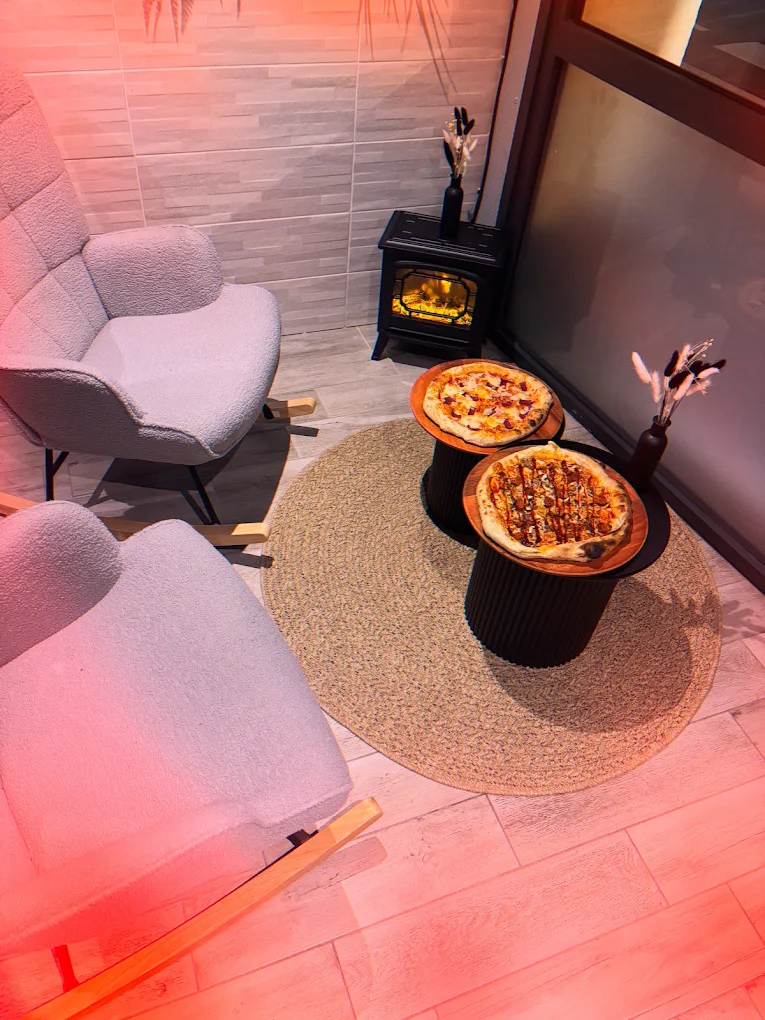 Coin salon de la salle : deux fauteuils en tissu bouclette, un tapis rond et un poêle, avec deux pizzas posées sur des planches